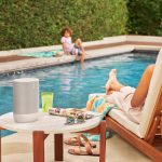 A mother is sitting by the pool while her child soaks their feet in the water on the other side. Next to the mother is a table with a Sonos wireless speaker.