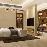 A modern and stylish bedroom with white bedding, nightstands, and a wooden shelving unit along the far wall. The room is illuminated by a modern chandelier and has a warm, neutral color palette.