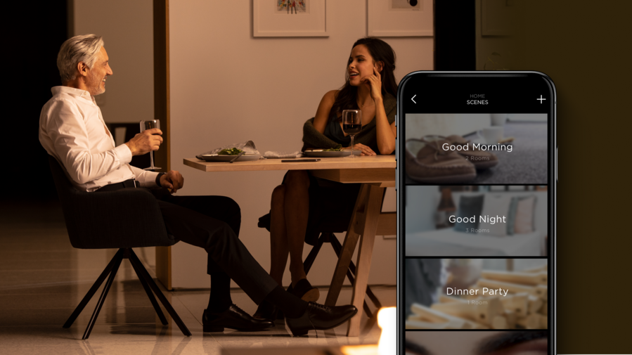 A couple has dinner in a warmly illuminated room while a phone shows different scenes in the Savant app.