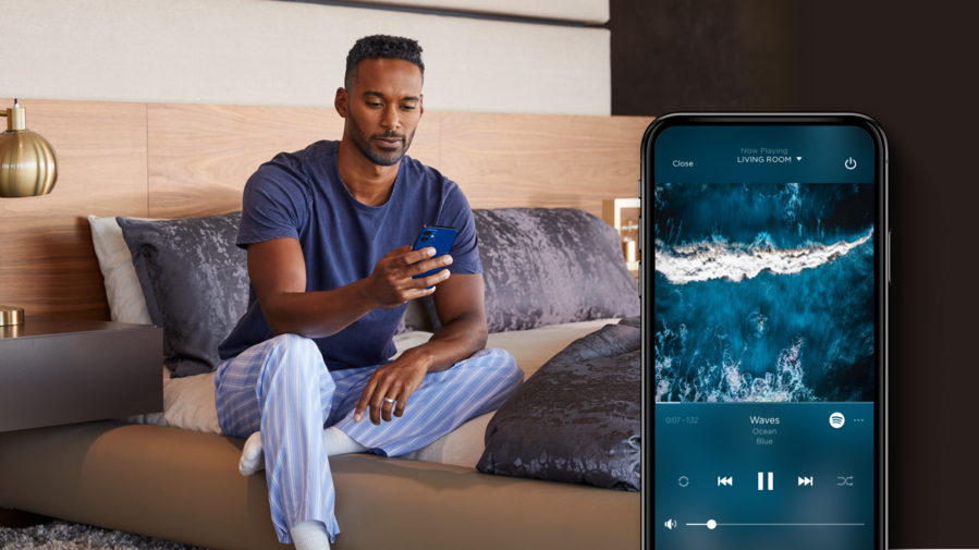 Man sitting on a modern bed, using a smartphone to control music playback, with a close-up of the phone screen showing a streaming app playing ocean sounds.