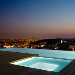 Modern rooftop infinity pool and hot tub overlooking a glowing city skyline at dusk, with soft ambient lighting enhancing the luxurious outdoor setting.